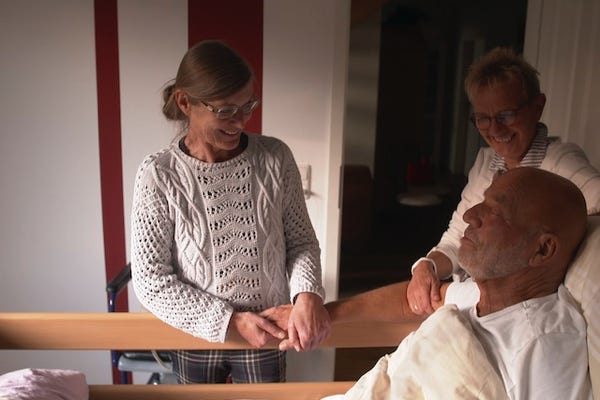 Mann sitzt in Bett, Palliativärztin und Begleitung